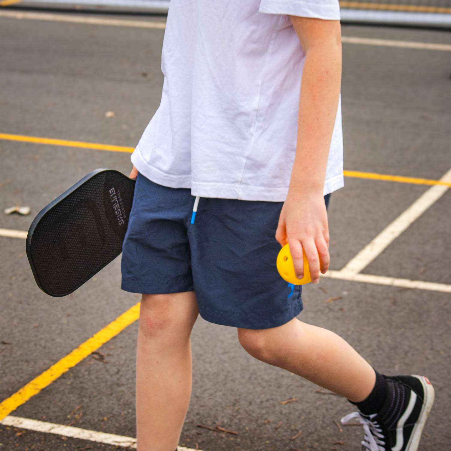 Pickleball Full Size Net Set, Action shot, kid holding a black polycore paddle and a yellow outdoor pickleball