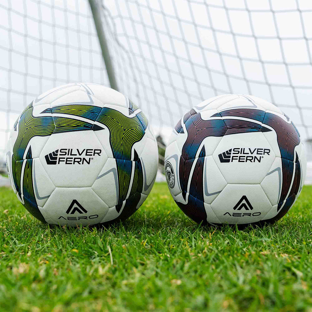 
Two Aero soccer match balls on a grass field in front of a goal net. The ball on the left is a size 5, while the ball on the right is a size 4, both displaying the Silver Fern logo.